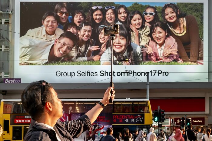 Street coincidences caught on camera: A person taking a selfie with a selfie stick, aligning perfectly with a large billboard of a group selfie.
