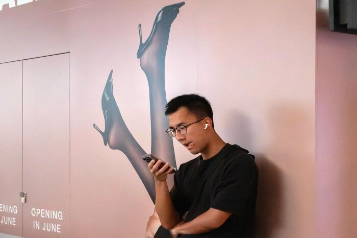 Street coincidences caught on camera: A man in glasses looking at his phone, aligning with a poster of high-heeled legs.