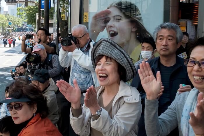 Crowd of people watching an event, with a woman's face on a billboard appearing to look over them, a street coincidence.