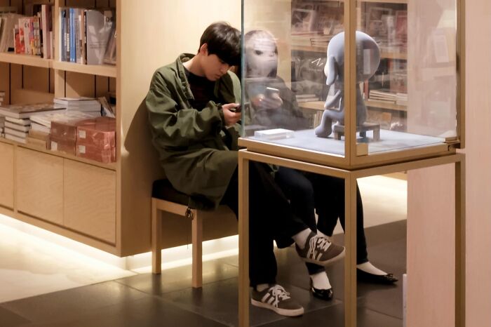 A man on his phone next to a glass display reflects his face onto a small statue's head, a street coincidence.