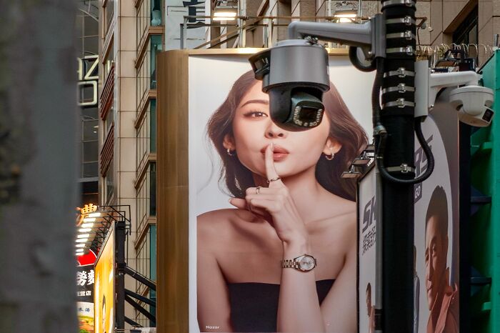 Street coincidences caught on camera: A CCTV camera lens aligns with a woman's finger on a billboard, creating a shushing pose.