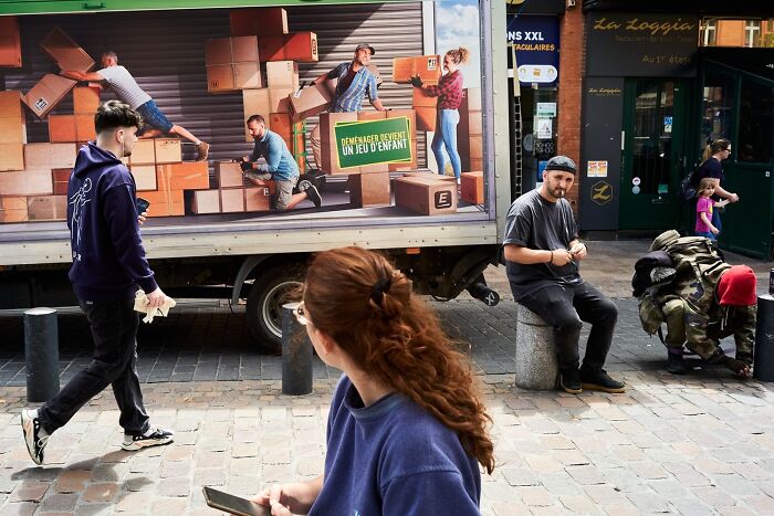 A perfectly timed street photo of people on a street with a truck mural depicting moving boxes, revealing an extraordinary moment in everyday life.