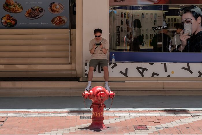 A man stands on a fire hydrant, looking at his phone, a street coincidence caught on camera.
