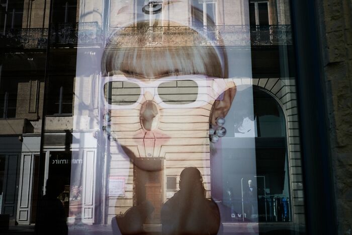 A perfectly timed street photo showing a reflection of a woman with sunglasses overlaying a building, revealing an extraordinary moment in everyday life.