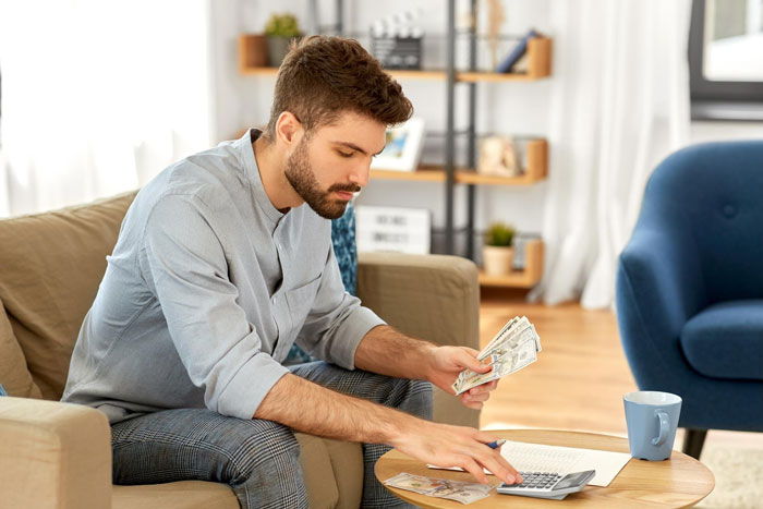 A guy counting money and using a calculator at home, managing inheritance for his sis. A guy counting money and using a calculator at home, managing inheritance for his sis.