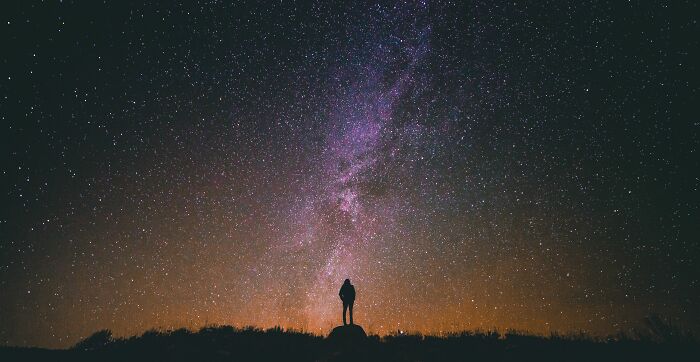 A person stands under a vibrant night sky full of stars, pondering deeply disturbing cults.