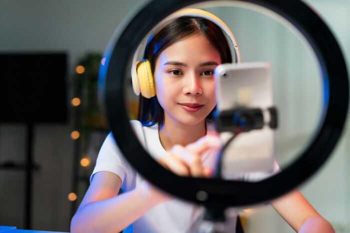 A young woman with headphones and a ring light, filming on her phone, potentially discussing internet rabbit holes or cults.