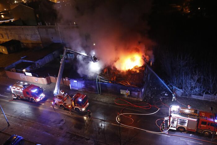 An aerial view of a building engulfed in a large fire at night, with multiple fire trucks and firefighters battling the blaze, showing deeply disturbed internet rabbit holes.