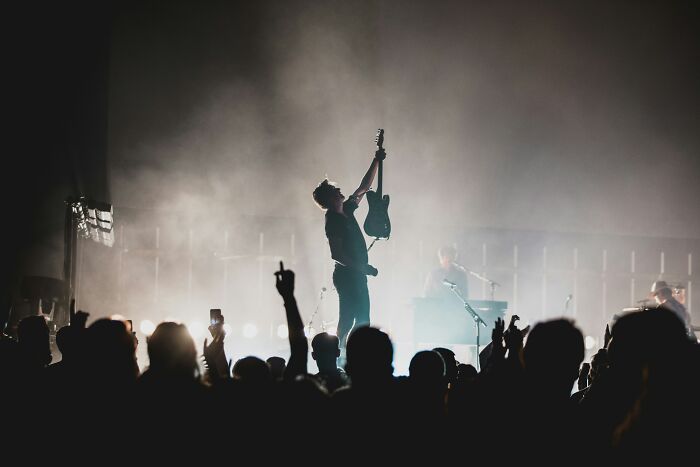 A musician holding a guitar high on stage with a crowd, contrasting with the dark themes of cults.