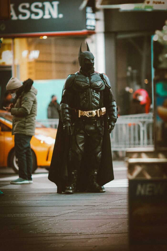 A person dressed as Batman in a black suit with a cape and golden belt buckle stands on a city street, featuring fascinating things about the world.