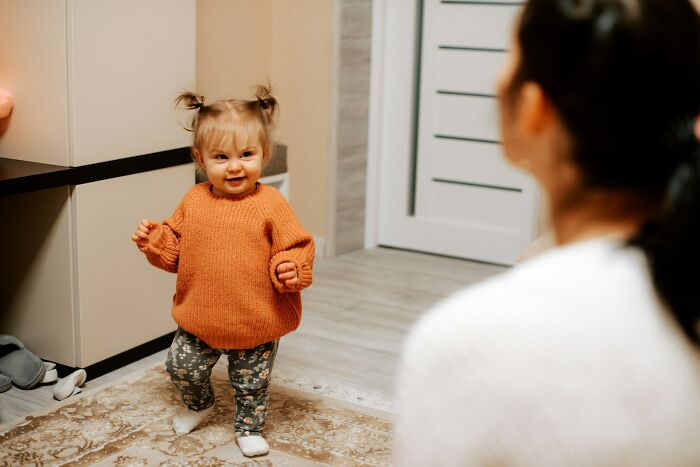 A happy toddler in an orange sweater walking towards an adult, perfect for babysitting stories.
