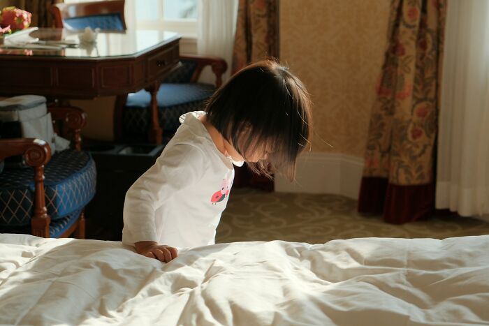 A young child in a white shirt peeking over a bed in a sunny room, representing innocent yet captivating babysitting stories.