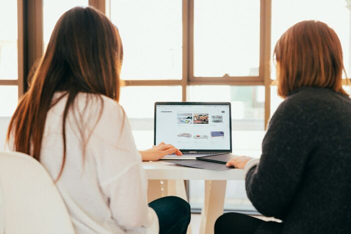 Two women seated, looking at a laptop screen displaying a product website, emphasizing micro feminism clap backs in a bright room.