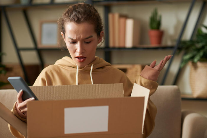 A woman looking shocked while holding her phone and opening a cardboard box, disappointed about a fake designer bag. A woman looking shocked while holding her phone and opening a cardboard box, disappointed about a fake designer bag.