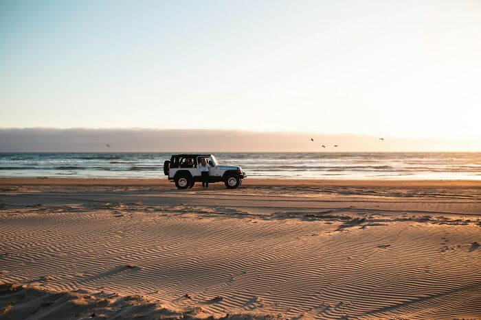 A white Jeep parked on a sandy beach next to the ocean, showcasing strange jobs for earning money.