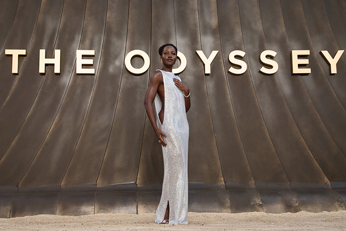 Lupita Nyong'o in a shimmering gown in front of THE ODYSSEY text. She is being roasted over a woke question.