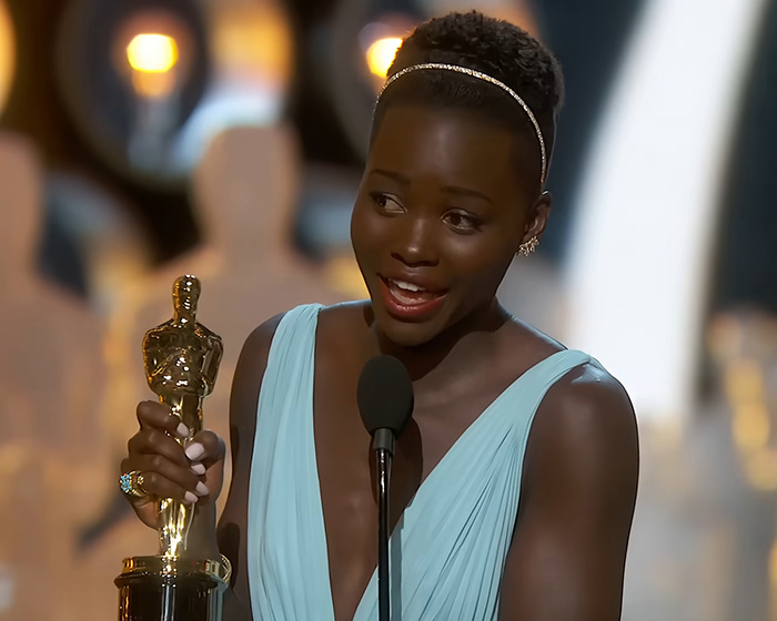 Lupita Nyong'o holds an Oscar trophy, smiling at an awards ceremony.