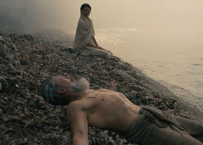 A scene from Odyssey with a man lying on the shore and a woman sitting nearby. Focuses on the woke question.