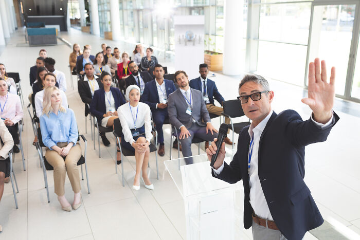 A speaker addresses workers at a conference, perhaps about corporate buzzwords and practices.