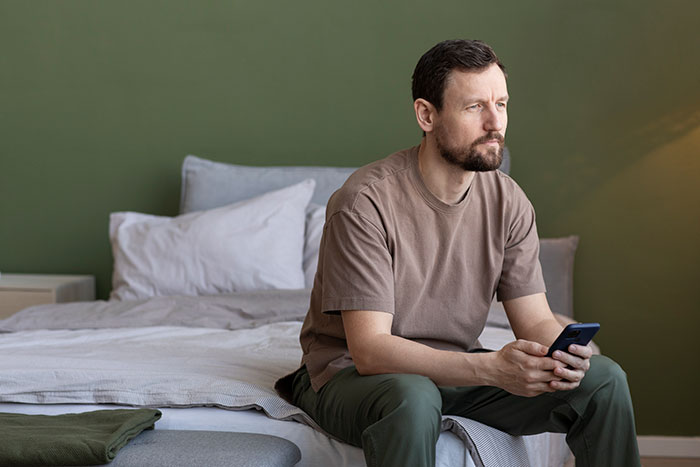 A man with a giant brain tumor sits on a bed, looking pensive, holding a phone, contemplating his second chance at life.
