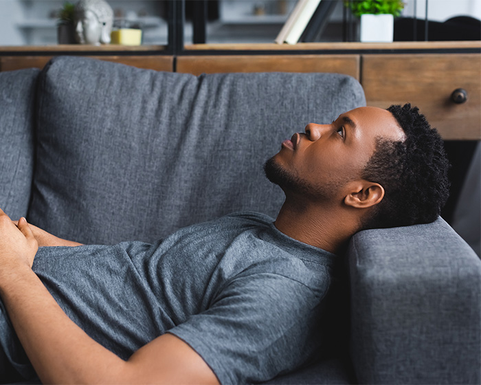 A lonely man lies on a gray sofa, looking upwards with a pensive expression, conveying feelings of loneliness. A lonely man lies on a gray sofa, looking upwards with a pensive expression, conveying feelings of loneliness.
