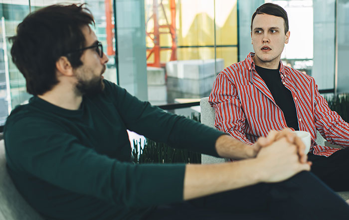 Two men in a modern setting, discussing the entitled man's plan to propose at his brother's wedding.