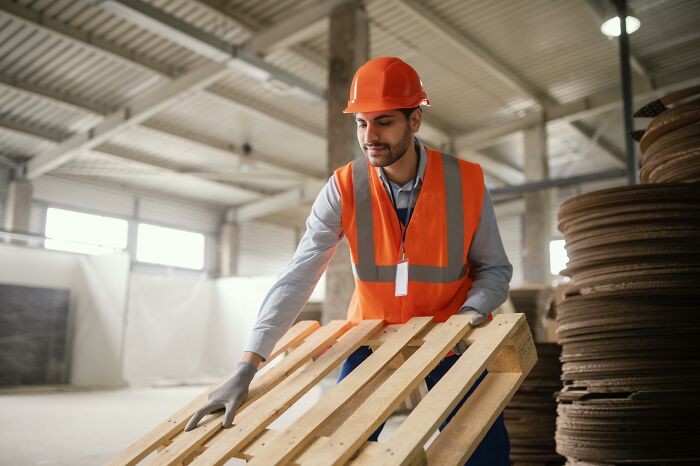 A worker in an orange vest and hard hat handles a wooden pallet, representing industries that make millions.