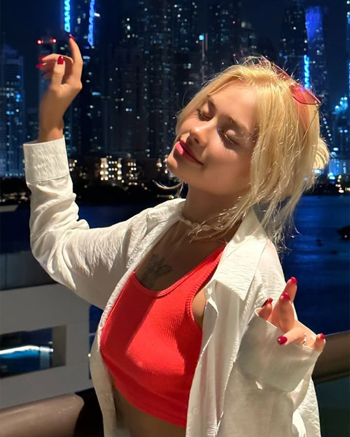 A famous model with blonde hair, smiling with closed eyes, wearing a white shirt and red top against a city nightscape. A famous model with blonde hair, smiling with closed eyes, wearing a white shirt and red top against a city nightscape.