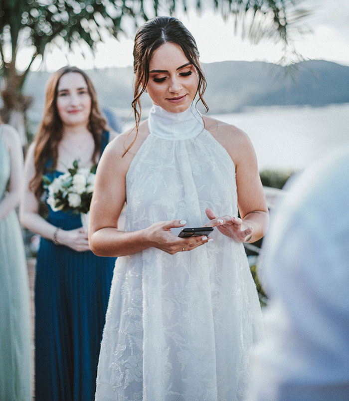 A bride in a white dress looking at her phone, with a bridesmaid in blue holding flowers in the background. This image depicts a bride distracted by her phone, a potential wedding red flag suggesting disinterest or preoccupation during her marriage.