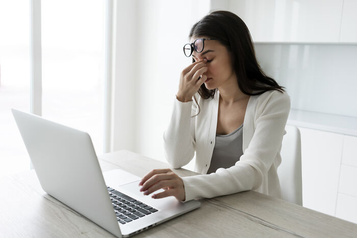 A woman with glasses rubs her eyes while looking at a laptop, showing stress from corporate buzzwords and practices.