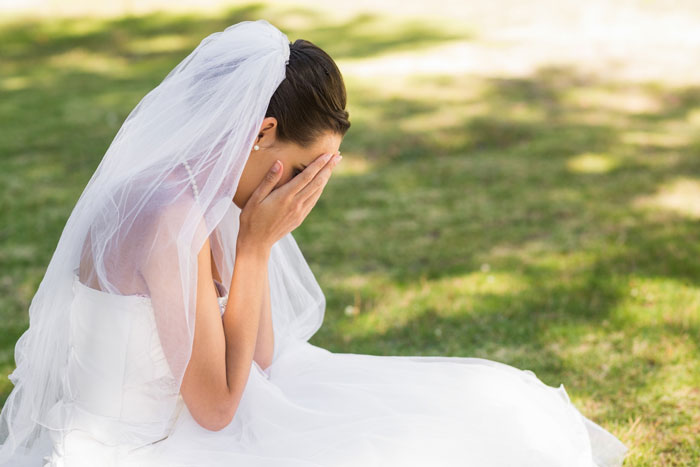 A frustrated bride in a white wedding dress sits on grass, covering her face with her hands, due to toxic MIL. A frustrated bride in a white wedding dress sits on grass, covering her face with her hands, due to toxic MIL.