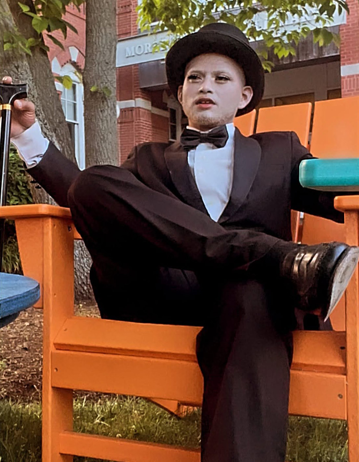 A mime in a black suit, bow tie, and hat, holding a cane and sitting outdoors in an orange chair. A mime in a black suit, bow tie, and hat, holding a cane and sitting outdoors in an orange chair.