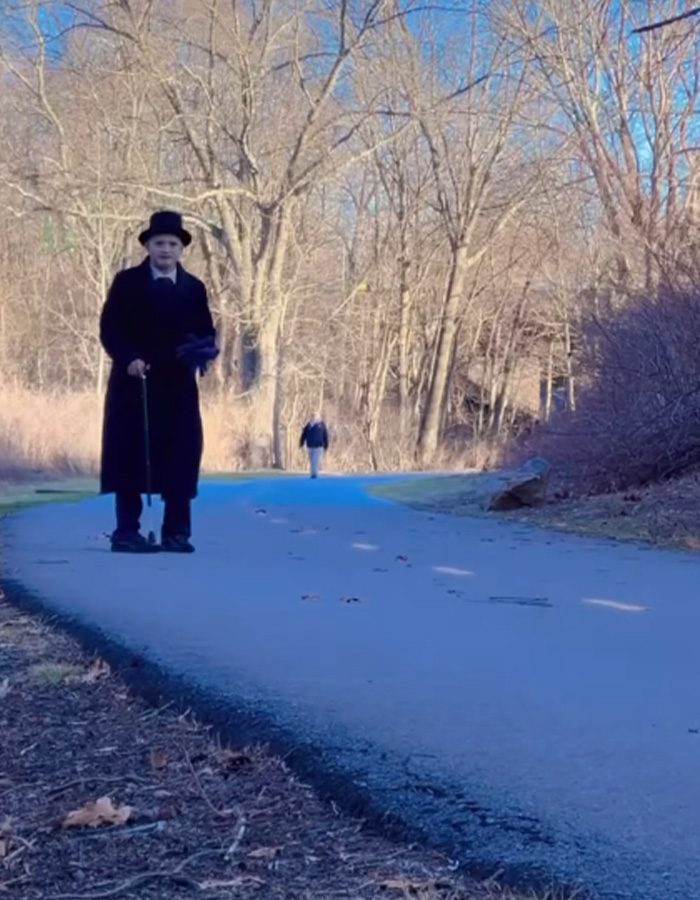 A man dressed as a mime, wearing a black coat and top hat, walking on a paved path, in a cooking oil stunt. A man dressed as a mime, wearing a black coat and top hat, walking on a paved path, in a cooking oil stunt.