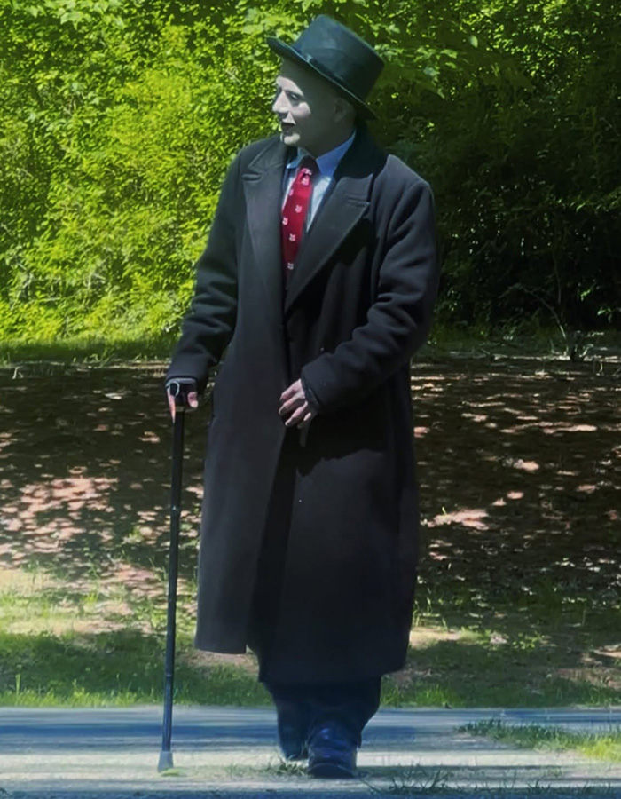 A mime accused of terrorizing women, dressed in a black coat and hat, holding a cane, with green foliage behind him. A mime accused of terrorizing women, dressed in a black coat and hat, holding a cane, with green foliage behind him.