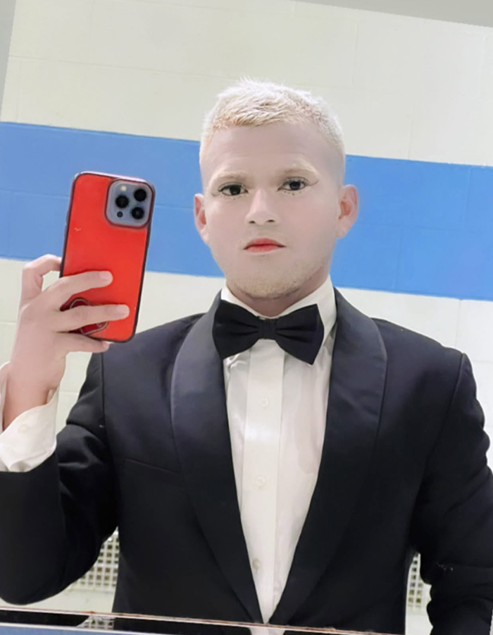 A man with pale makeup and blonde hair, dressed in a tuxedo, holding a red phone, like a mime accused of terrorizing women. A man with pale makeup and blonde hair, dressed in a tuxedo, holding a red phone, like a mime accused of terrorizing women.