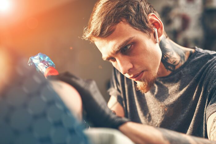 A focused tattoo artist with a beard and neck tattoos, carefully working on a client's permanent tattoo, avoiding spelling errors.