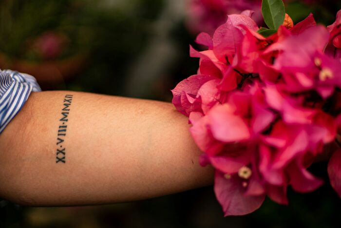 An arm with a Roman numeral tattoo, juxtaposed with pink flowers, illustrating potential tattoo spelling errors.