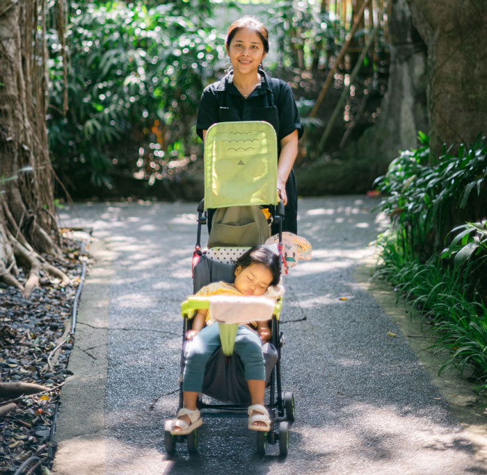 A nanny pushing a sleeping child in a stroller during a family vacation, walking on a shaded path.