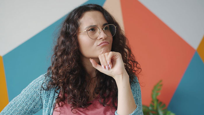 A thoughtful woman with curly hair and glasses, pondering a decision about her family vacation and nanny.