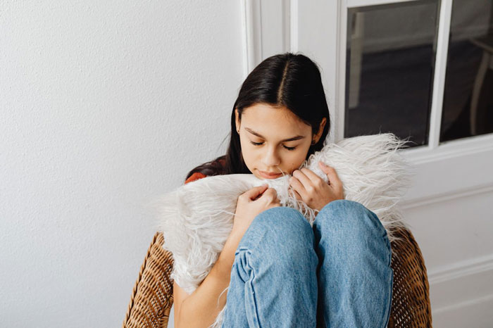 A woman on a family vacation looks down sadly, hugging a fluffy white pillow, contemplating the nanny decision.