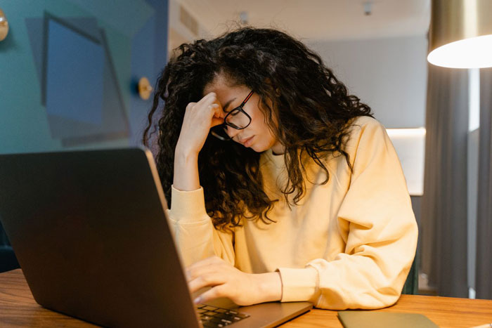 A woman looking stressed, holding her head, and working on a laptop, related to a family vacation decision.