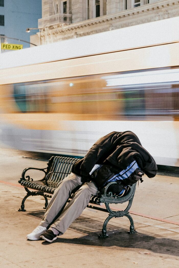 A person sleeping on a park bench at night with a blurred bus passing, showing a disturbing public sight.