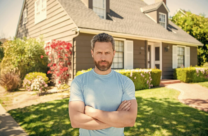 A man with a serious expression in front of his house, standing as if protecting his garden from an entitled neighbor.