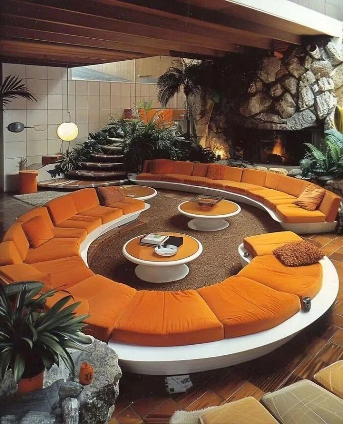 An iconic furniture design with a large, curved orange sofa in a sunken living area with a stone fireplace and tropical plants.