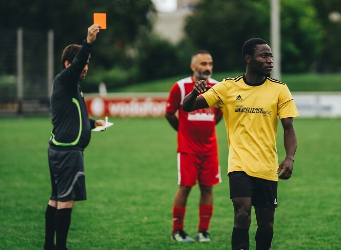 A soccer referee holding up a red card to a player in a yellow jersey on a green field. Fascinating things about the world.