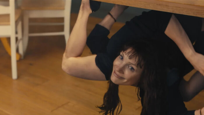 A woman with red hair smiles from under a wooden table, from The Odyssey trailer. A woman with red hair smiles from under a wooden table, from The Odyssey trailer.