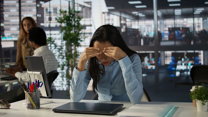 A stressed worker in an office rubs her temples, overwhelmed by corporate buzzwords and practices.