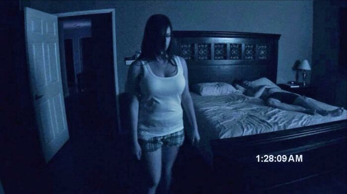 A woman standing in a dimly lit bedroom while a man sleeps, a terrifying scene from horror movies.