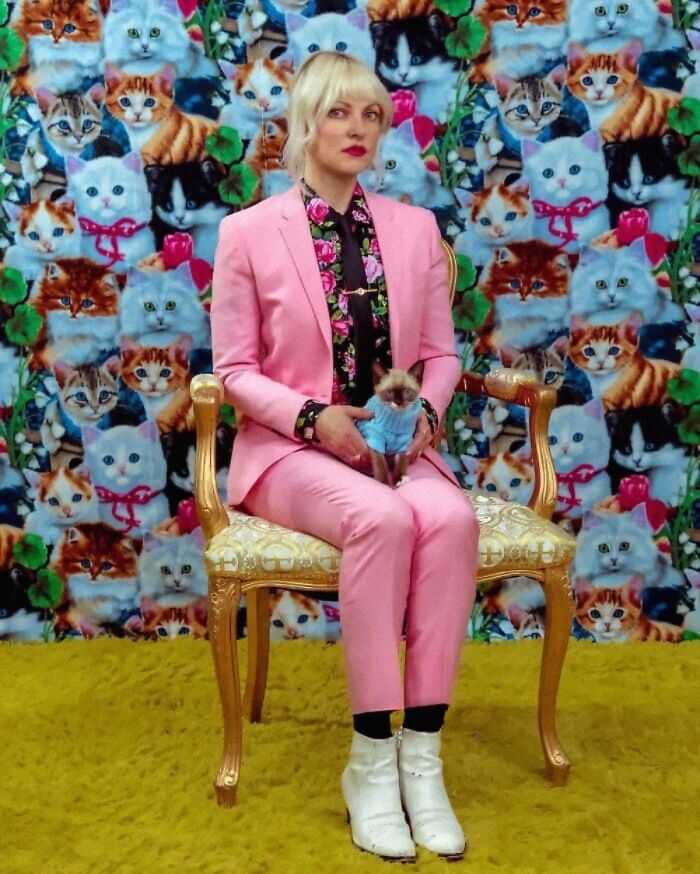 A woman in a pink suit sitting on a gold chair, holding a small dog in front of a wall of cat photos.