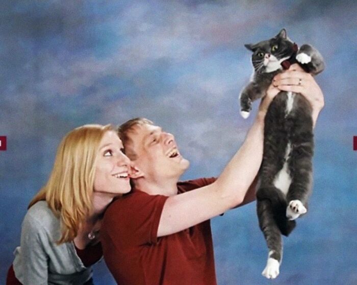 A laughing couple holding up their cat against a blue backdrop, capturing a funny and adorable animal photo.